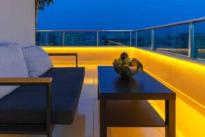A cozy balcony features a dark couch and a sleek table with a bowl of colorful fruits, illuminated by warm LED lights at dusk.