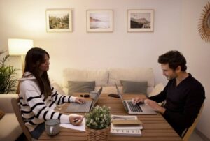 Two individuals work on laptops at a wooden table, surrounded by a cozy living space with plants and framed pictures on the wall.