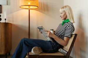 A woman in a striped shirt and green bandana sits casually in a chair, reading a magazine beside a stylish lamp.