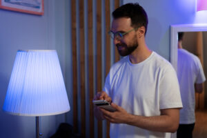 A person in a white t-shirt is standing by a blue-lit lamp, holding a smartphone in one hand and looking at the screen.
