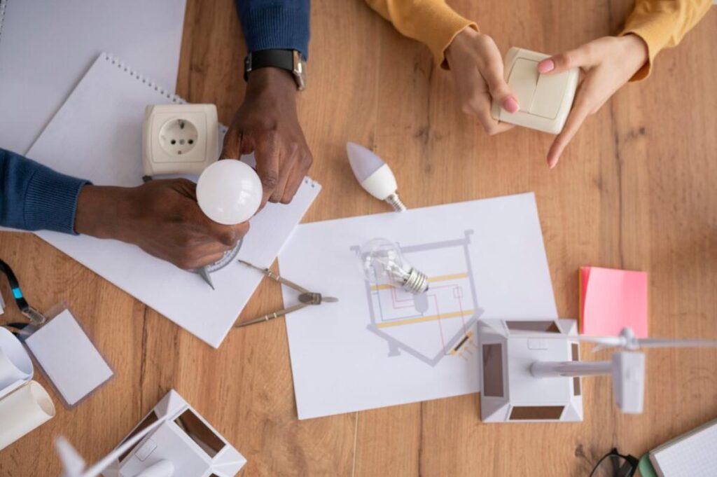 Two hands working together with a light bulb, electrical socket, and blueprints on a wooden table, focused on a lighting project.