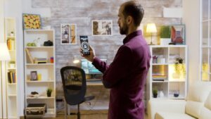 A man in a maroon shirt uses a smartphone to control smart lights in a modern, stylish room with shelves and a desk.