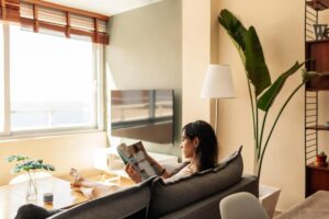 A person sits on a gray sofa reading a magazine, with a large TV and a tall plant in a bright, stylish living room.
