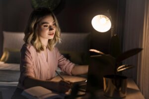 A person in a soft pink shirt writes in a notebook at a desk illuminated by a warm lamp, with a plant nearby.