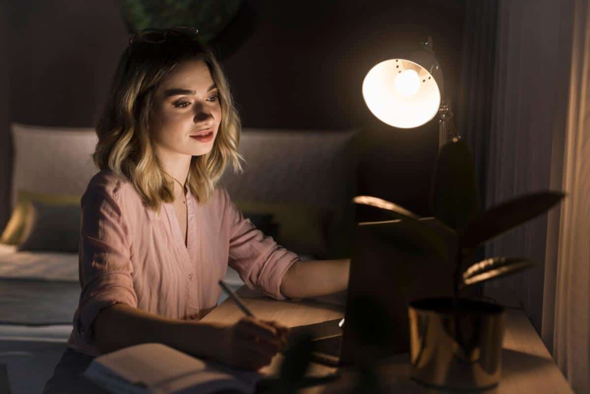 A person in a soft pink shirt writes in a notebook at a desk illuminated by a warm lamp, with a plant nearby.