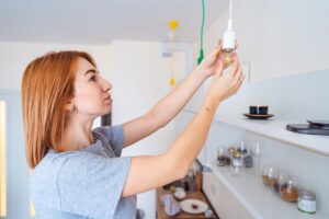A person with red hair reaches up to install a light bulb in a modern kitchen with minimalist decor and various kitchenware on shelves.