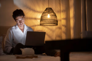 A person sitting on a bed, working on a laptop, with a warm glow from a hanging lamp and soft shadows in a cozy bedroom setting.