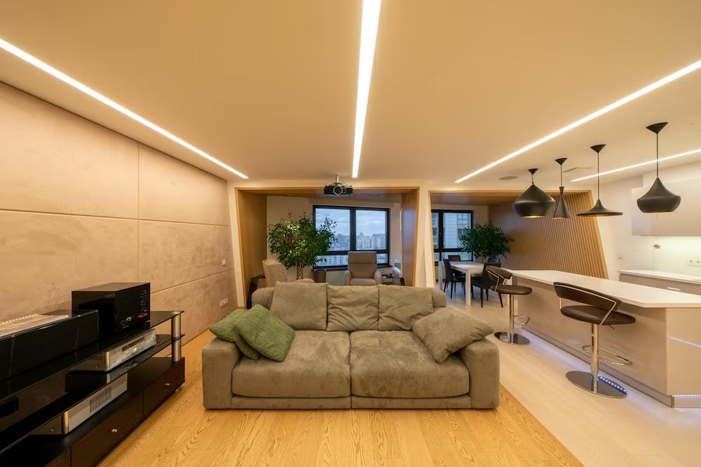 Modern living room with a plush sofa, sleek bar stools, and minimalist decor, featuring plants and ambient lighting. 
