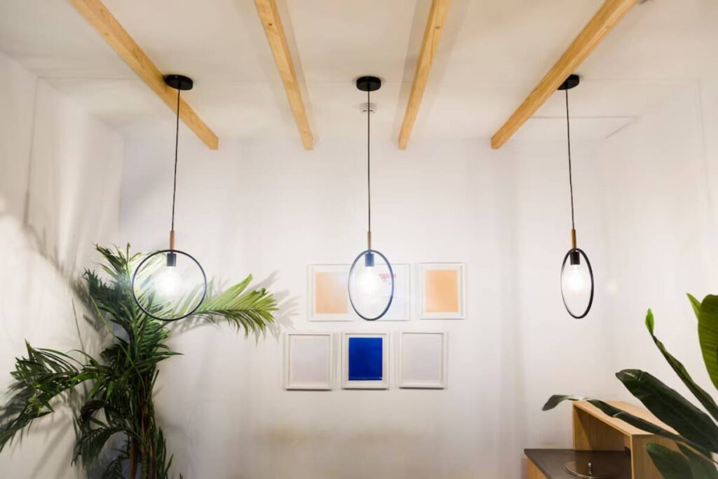 Three stylish pendant lights hang from wooden beams in a bright room, complemented by a green plant and framed artwork on the wall.