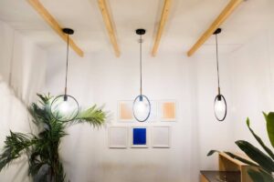Three stylish pendant lights hang from wooden beams in a bright room, complemented by a green plant and framed artwork on the wall.