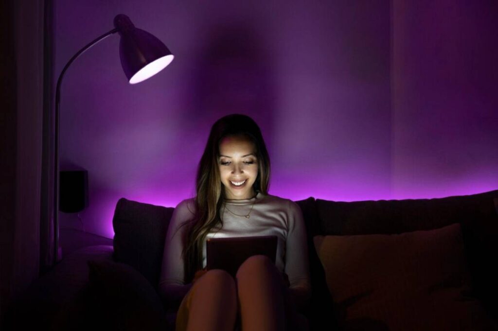 A person sitting on a couch, illuminated by a warm lamp and purple LED lights, holding a tablet in a cozy setting.