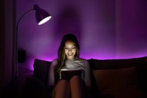 A person sitting on a couch, illuminated by a warm lamp and purple LED lights, holding a tablet in a cozy setting.