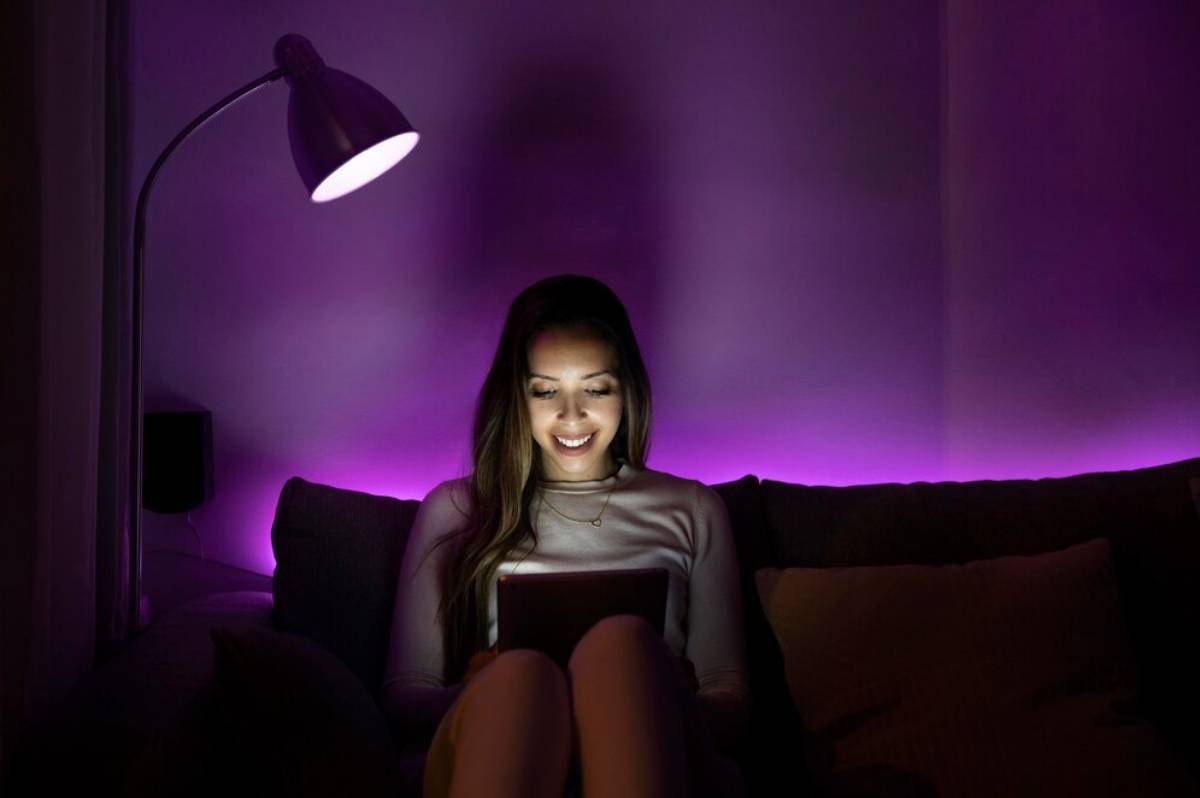 A person sitting on a couch, illuminated by a warm lamp and purple LED lights, holding a tablet in a cozy setting.